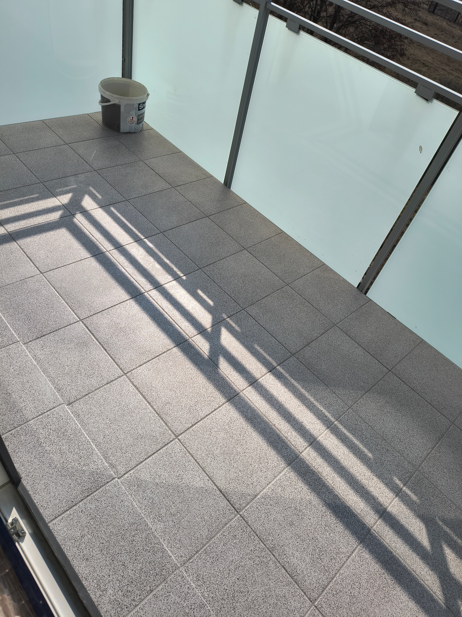 Wykończony balkon z szarymi płytkami podłogowymi i szklaną balustradą. Wiadro z farbą w rogu. Cień rzucany przez konstrukcję balkonu.