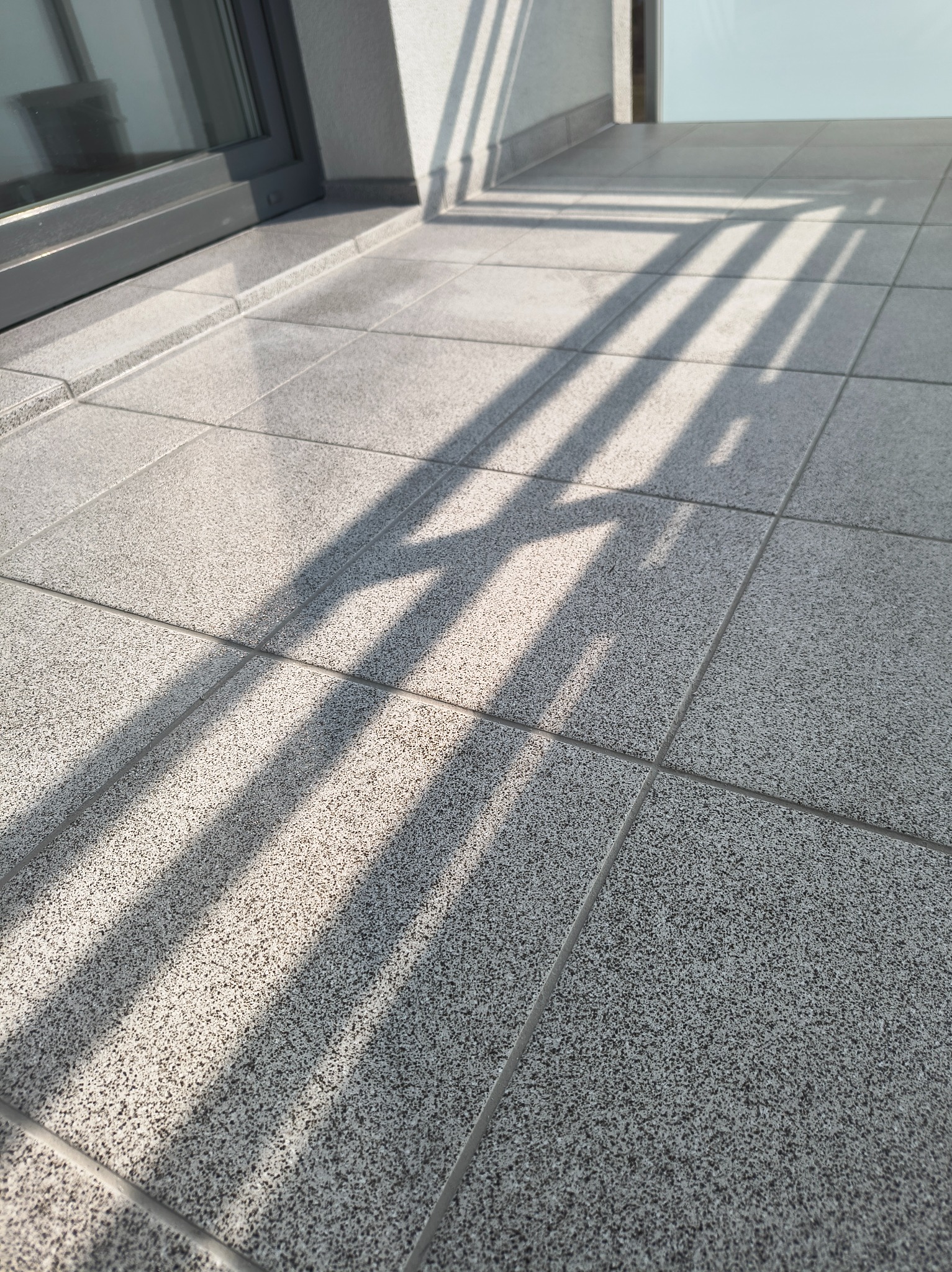 Jasnoszara, gładka podłoga z płytek na balkonie w słoneczny dzień. Widoczne cienie rzucane przez balustradę. Minimalistyczny design.