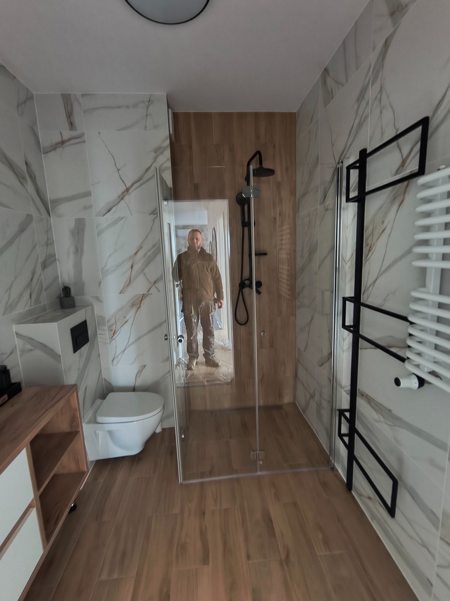 Wykończona łazienka z kabiną prysznicową typu walk-in, płytkami imitującymi marmur i drewno, oraz czarnymi akcentami. W lustrze odbija się osoba.