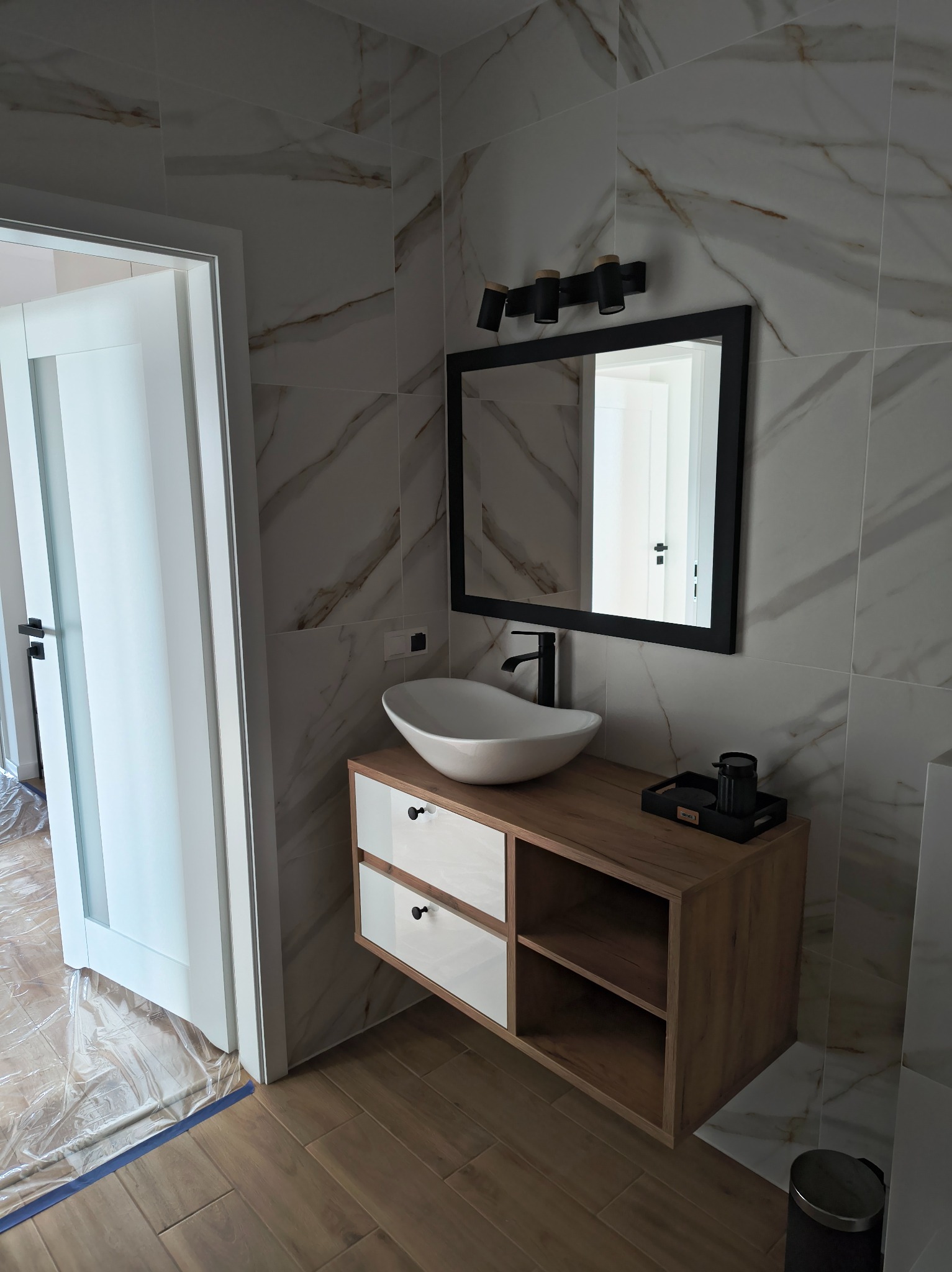 Nowoczesna łazienka po remoncie: biała umywalka nablatowa, szafka z drewnianym blatem i białymi frontami, lustro w czarnej ramie, marmurowe płytki na ścianach.