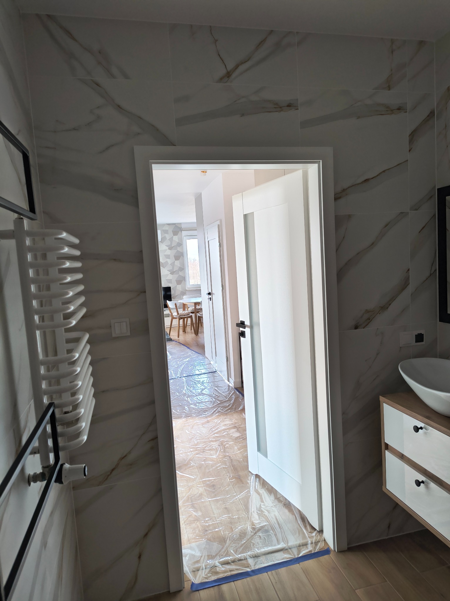 Wykończona łazienka z marmurowymi płytkami i białą armaturą, widok przez otwarte drzwi na zabezpieczoną folią podłogę w pokoju. Nowoczesny grzejnik i umywalka.