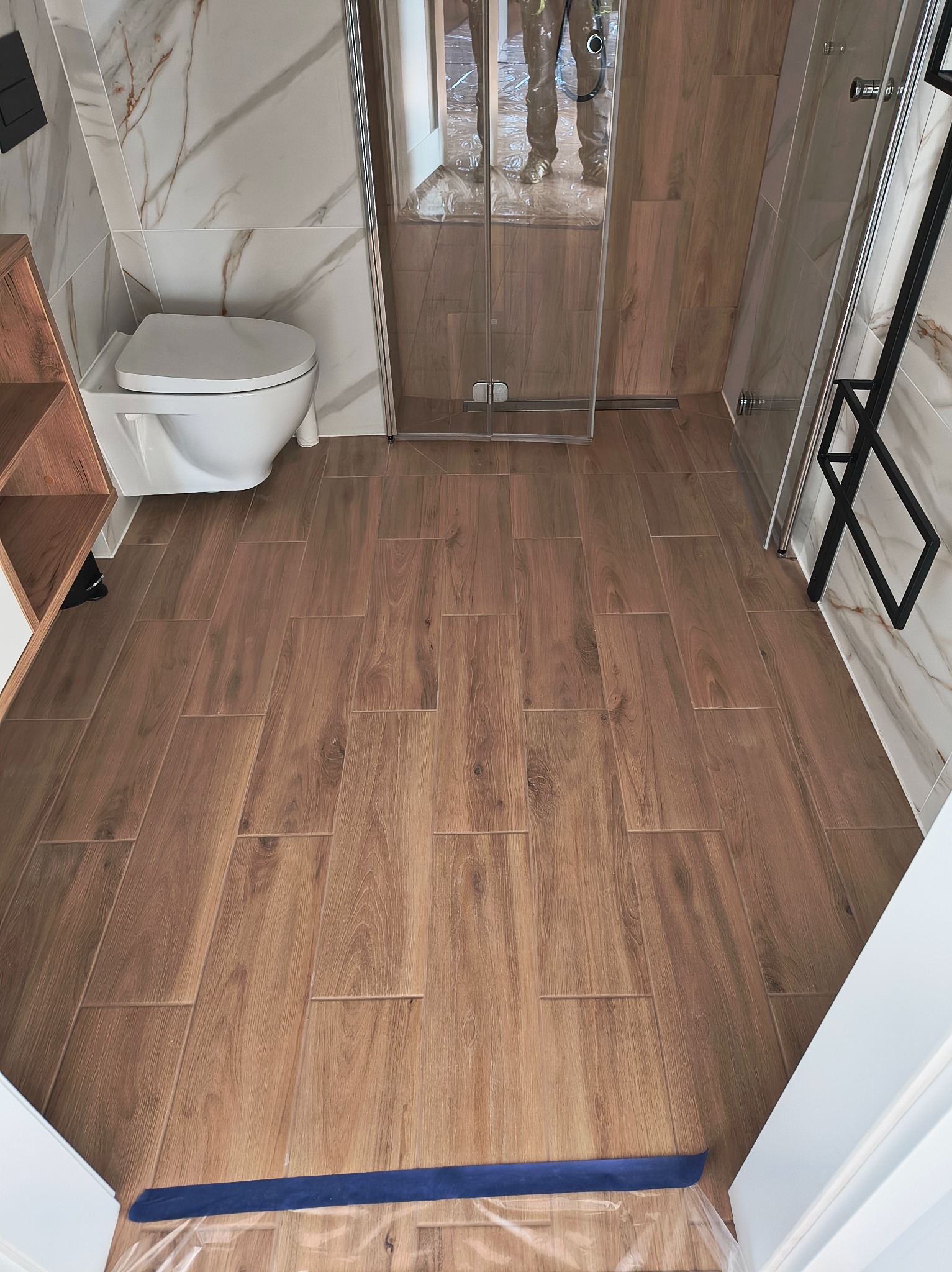 Ukończona łazienka z drewnopodobnymi płytkami podłogowymi, marmurowymi płytkami ściennymi i kabiną prysznicową. Widoczna toaleta i fragment regału.