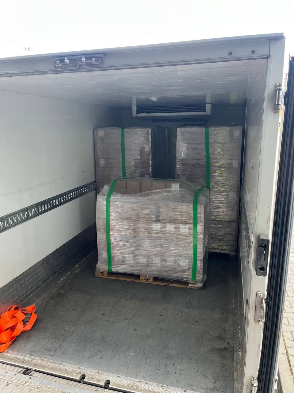 Wnętrze chłodni transportowej z paletami zabezpieczonymi folią i taśmami. Pomarańczowe pasy transportowe leżą na podłodze. Widoczny agregat chłodniczy.