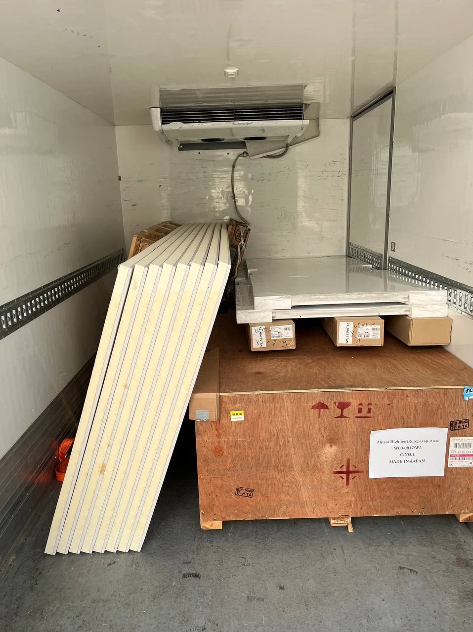 Wnętrze chłodni transportowej z ładunkiem: płyty izolacyjne oparte o ścianę, drewniana skrzynia z napisem 'MADE IN JAPAN', kartony. Widoczny agregat chłodniczy.