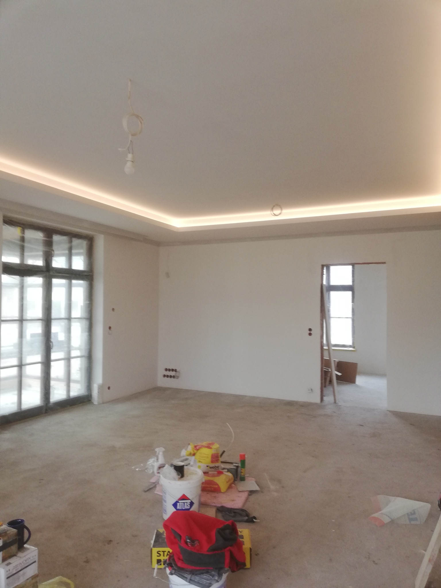 Wnętrze w trakcie prac: surowy beton na podłodze, ściany przygotowane do malowania, widoczne materiały budowlane i narzędzia. Oświetlenie sufitowe LED.