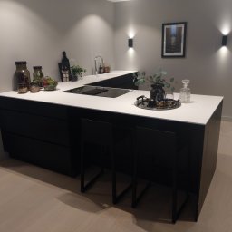 Nowoczesna kuchnia z czarnymi szafkami i białym blatem, oświetlona punktowymi lampami ściennymi. Na blacie dekoracje: wazon, taca z kieliszkami i ozdobne słoiki.