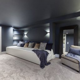 Eleganckie wnętrze: ciemnoszare ściany, białe drzwi i sofa z poduszkami. Lustro w czarnej ramie, fotel z narzutą i roślina doniczkowa dopełniają wystrój.