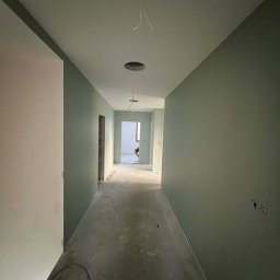 AG Wykończenia Wnętrz - Korytarz w odcieniach zieleni po świeżym malowaniu. Widoczne otwory na oświetlenie w suficie i gniazdka elektryczne na ścianie. Podłoga surowa, przygotowana do dalszych prac.