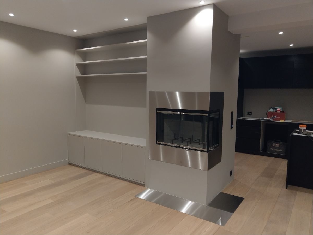 Minimalistyczny salon z zabudową meblową, kominkiem w obudowie ze stali nierdzewnej i jasną drewnianą podłogą. Nowoczesne oświetlenie punktowe.