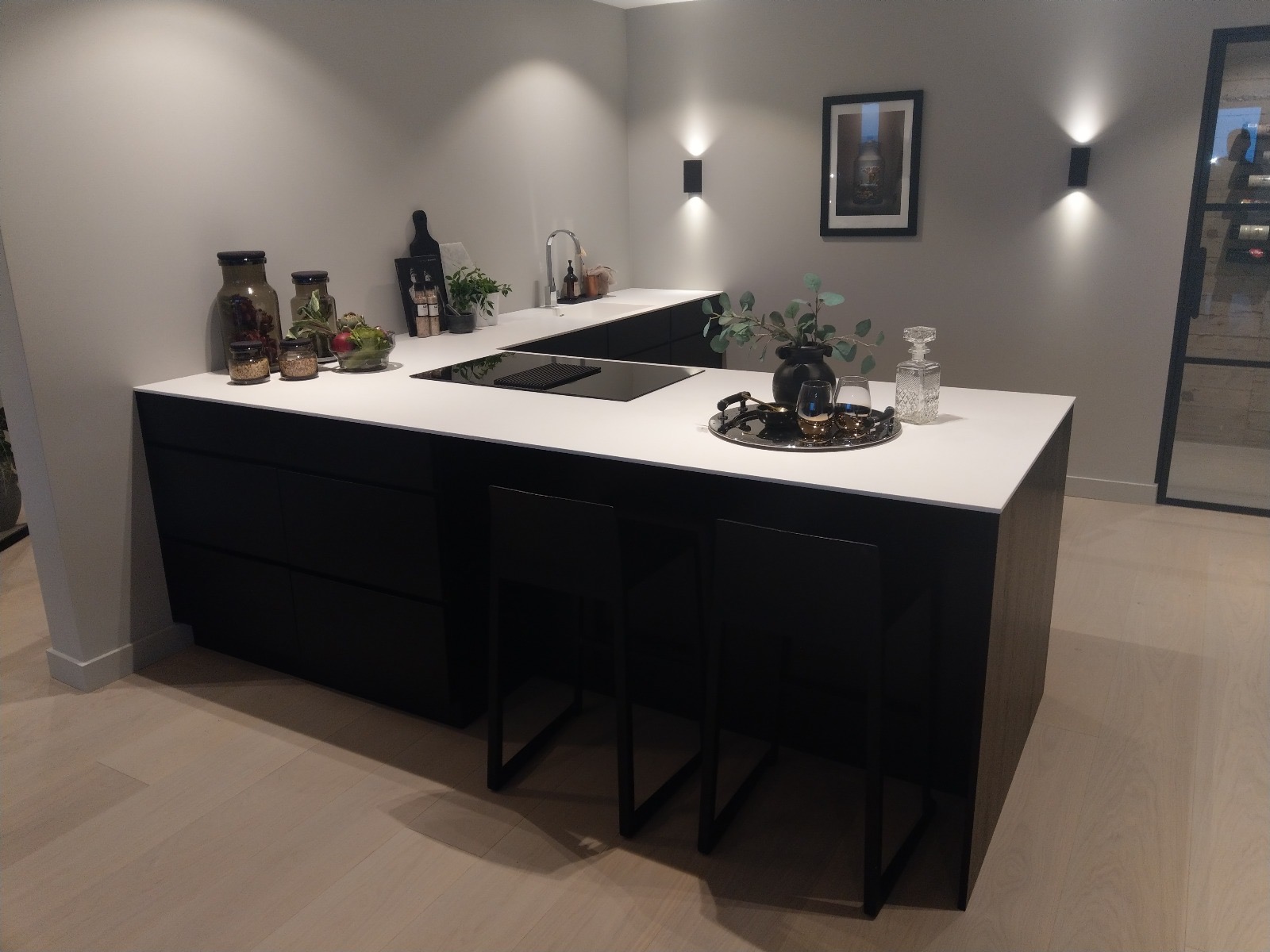 Nowoczesna kuchnia z czarnymi szafkami i białym blatem, oświetlona punktowymi lampami ściennymi. Na blacie dekoracje: wazon, taca z kieliszkami i ozdobne słoiki.