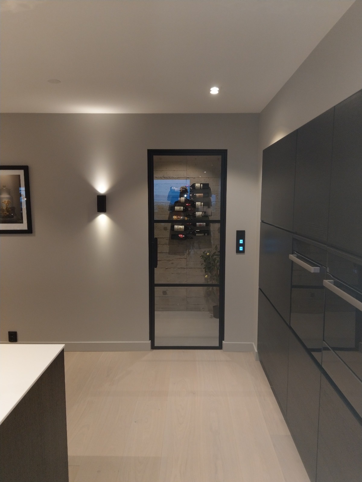 Nowoczesna kuchnia z czarną, przeszkloną ścianką działową, za którą widoczny regał na wino. Minimalistyczne oświetlenie i jasna podłoga podkreślają elegancki design.