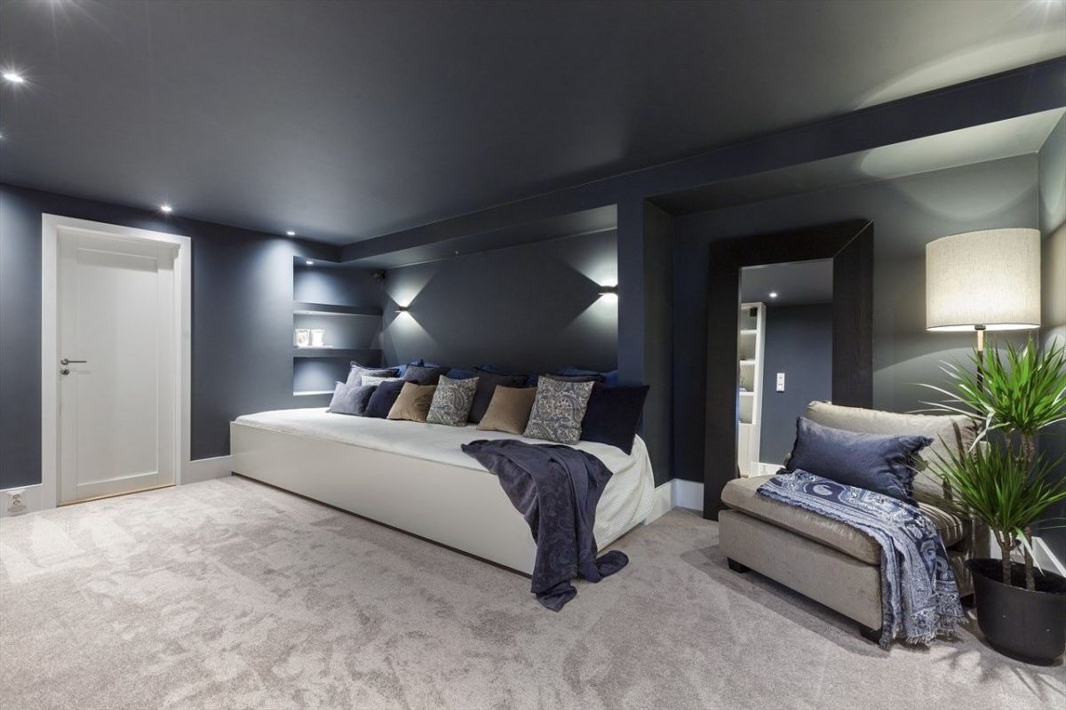 Eleganckie wnętrze: ciemnoszare ściany, białe drzwi i sofa z poduszkami. Lustro w czarnej ramie, fotel z narzutą i roślina doniczkowa dopełniają wystrój.