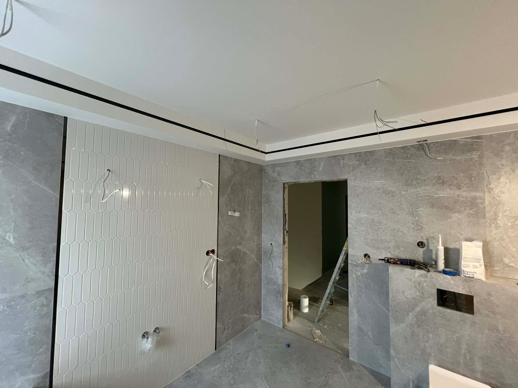 Wnętrze łazienki w trakcie wykańczania: szare płytki imitujące marmur, biała ściana z dekoracyjnymi płytkami, widoczne narzędzia i materiały budowlane, drabina w tle.