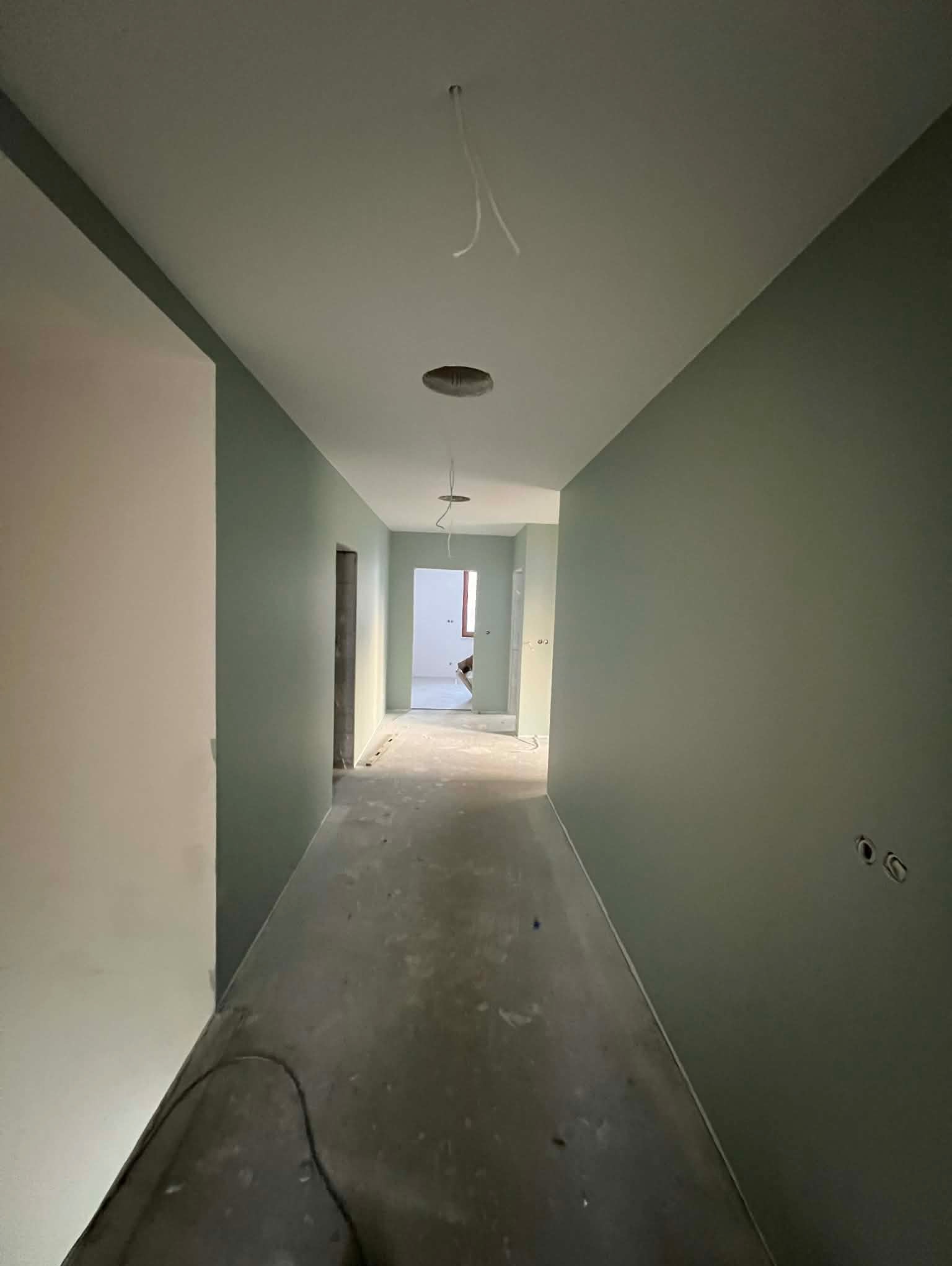 Korytarz w odcieniach zieleni po świeżym malowaniu. Widoczne otwory na oświetlenie w suficie i gniazdka elektryczne na ścianie. Podłoga surowa, przygotowana do dalszych prac.