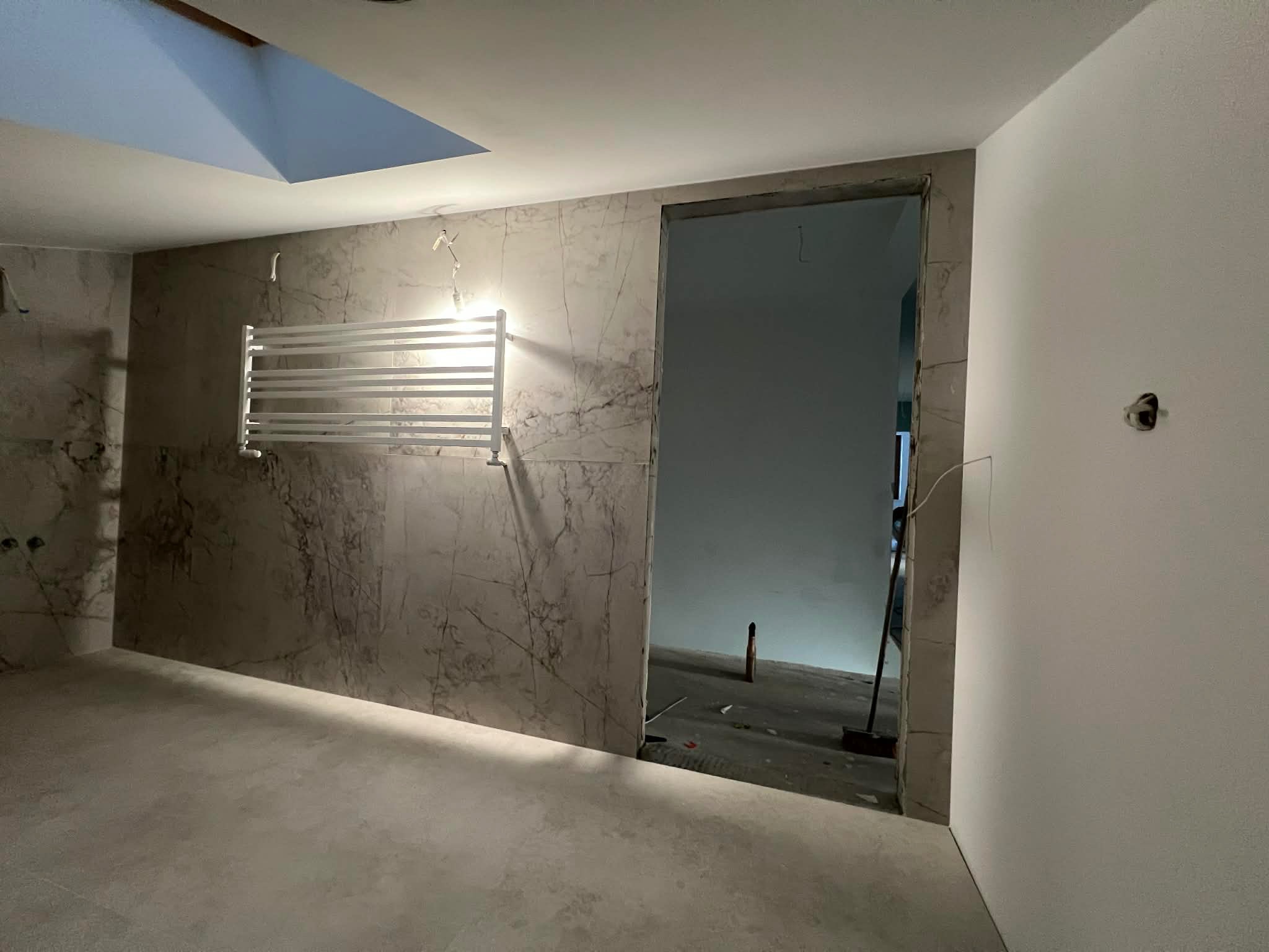 Wykończona łazienka z marmurowymi płytkami, grzejnikiem drabinkowym i otwartym przejściem do kolejnego pomieszczenia. Widoczne surowe wykończenie.