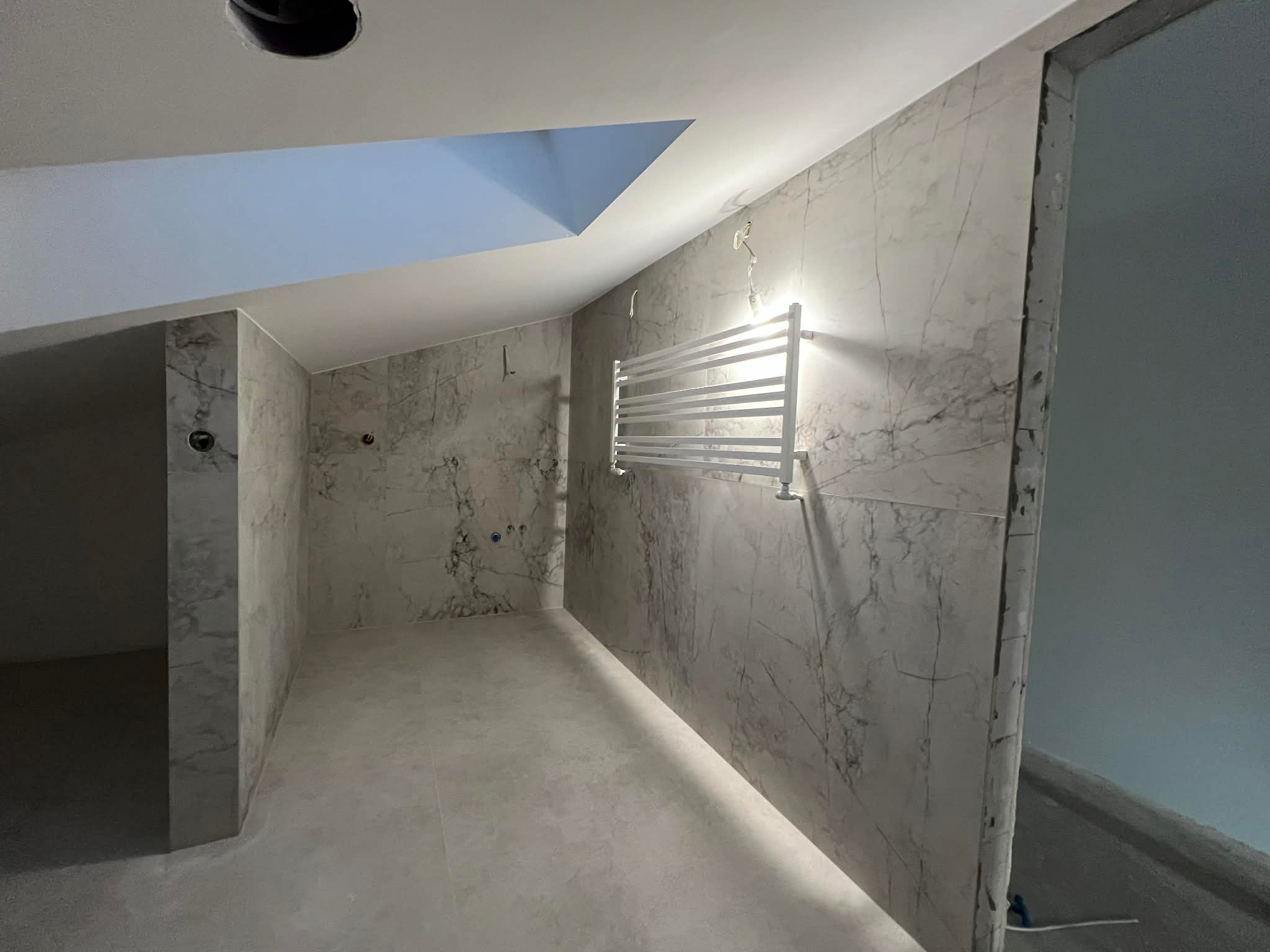 Wykończona łazienka na poddaszu z marmurowymi płytkami, grzejnikiem drabinkowym i oknem dachowym. Widoczne niedokończone obramowanie drzwi.