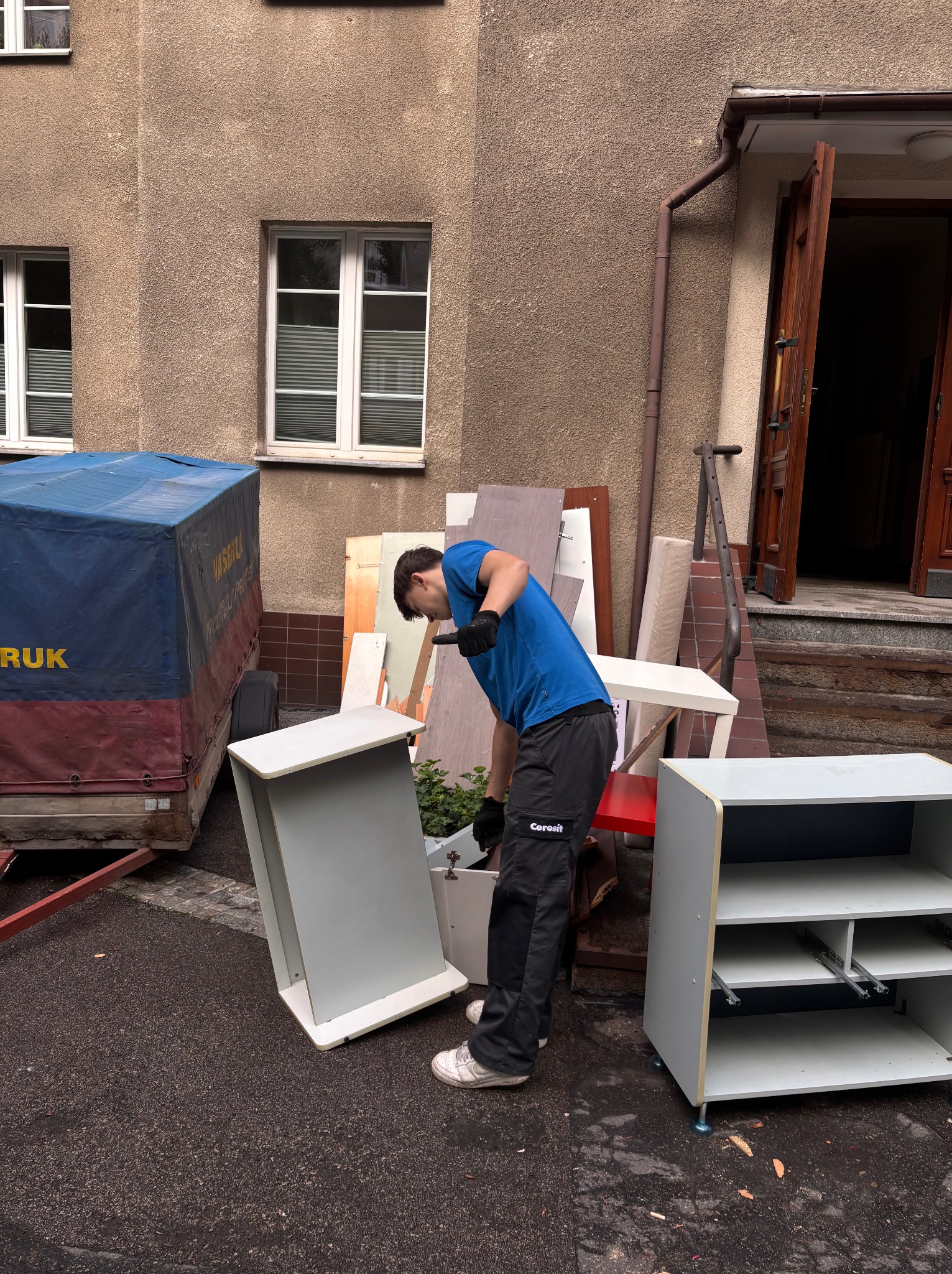 Mężczyzna w niebieskiej koszulce i czarnych spodniach wynosi meble z budynku. W tle przyczepka i otwarte drzwi wejściowe. Opróżnianie mieszkania.