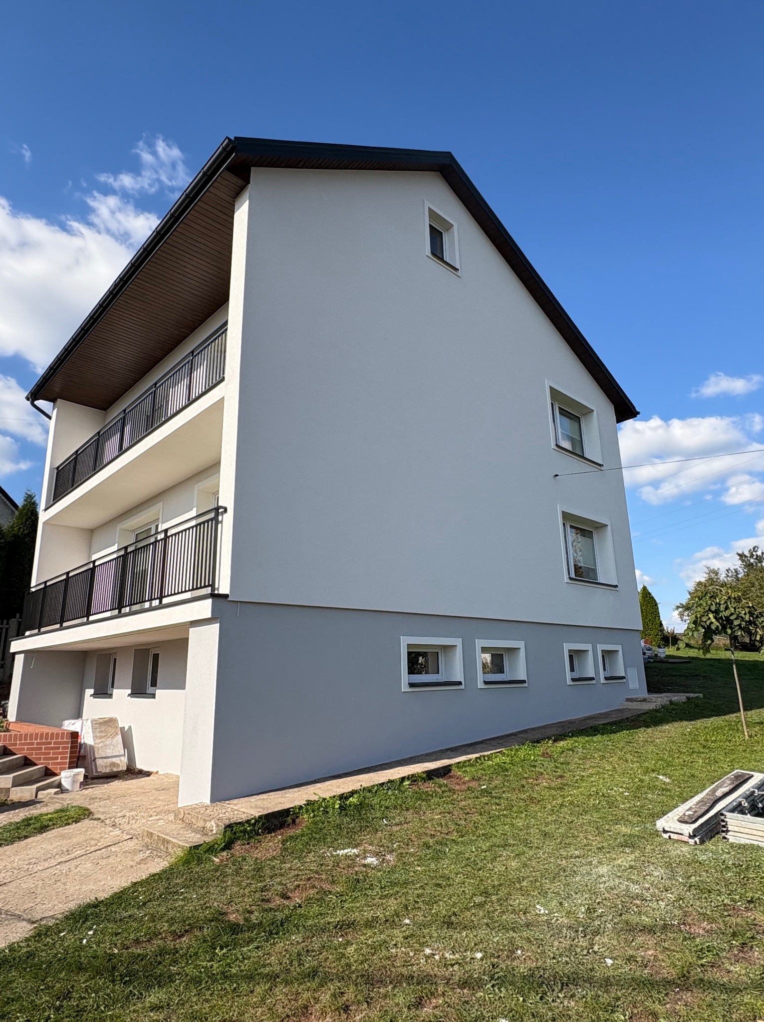 Dwukondygnacyjny dom po remoncie elewacji, z balkonami i szarą podmurówką, na tle błękitnego nieba. Widoczne schody wejściowe i fragment trawnika.