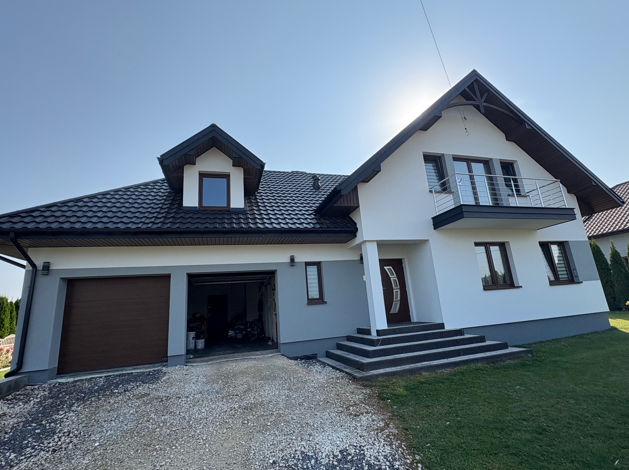 Dwukondygnacyjny dom z garażem i balkonem, wykończony w bieli i szarości, z ciemnobrązową stolarką okienną i dachem. Widok na elewację frontową w słoneczny dzień.