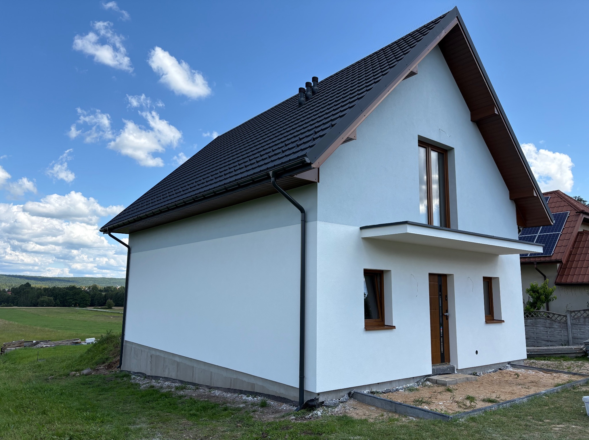 Nowoczesny dom jednorodzinny z białą elewacją i ciemną dachówką, widoczny z perspektywy ogrodu w słoneczny dzień. Minimalistyczny styl, drewniane okna i drzwi.