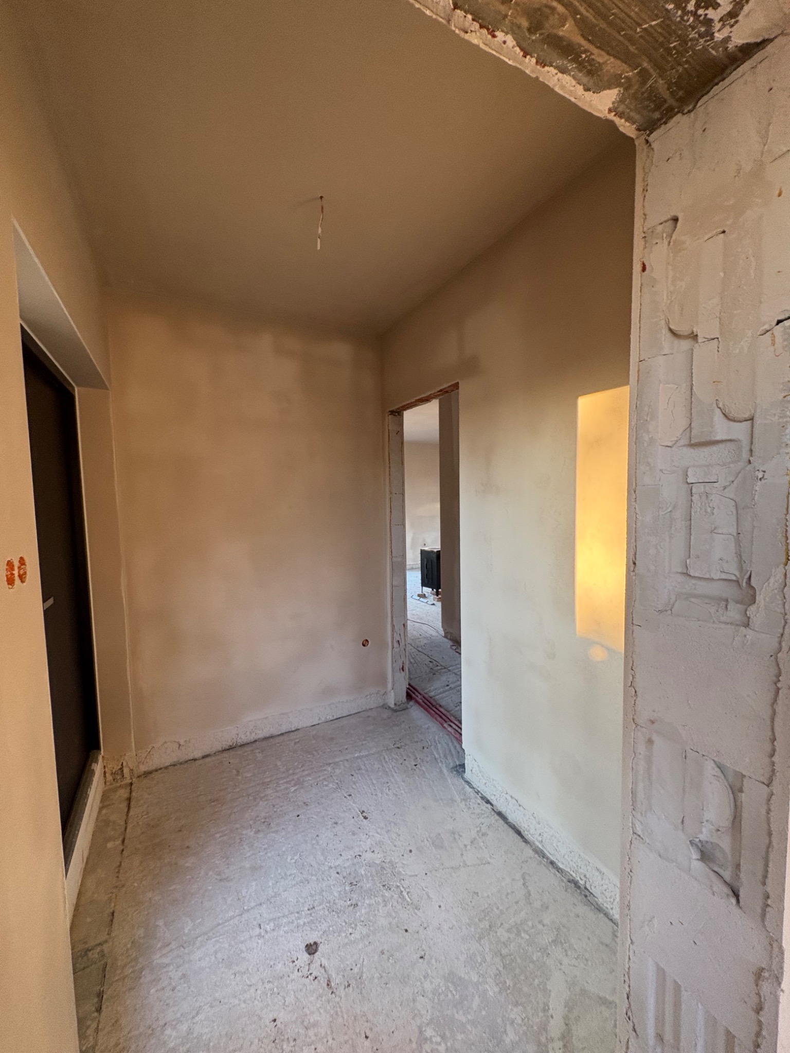 Wnętrze w trakcie remontu: ściany pokryte gładzią, surowy beton na podłodze, widoczne ślady po instalacjach. Otwór drzwiowy z surowym wykończeniem.