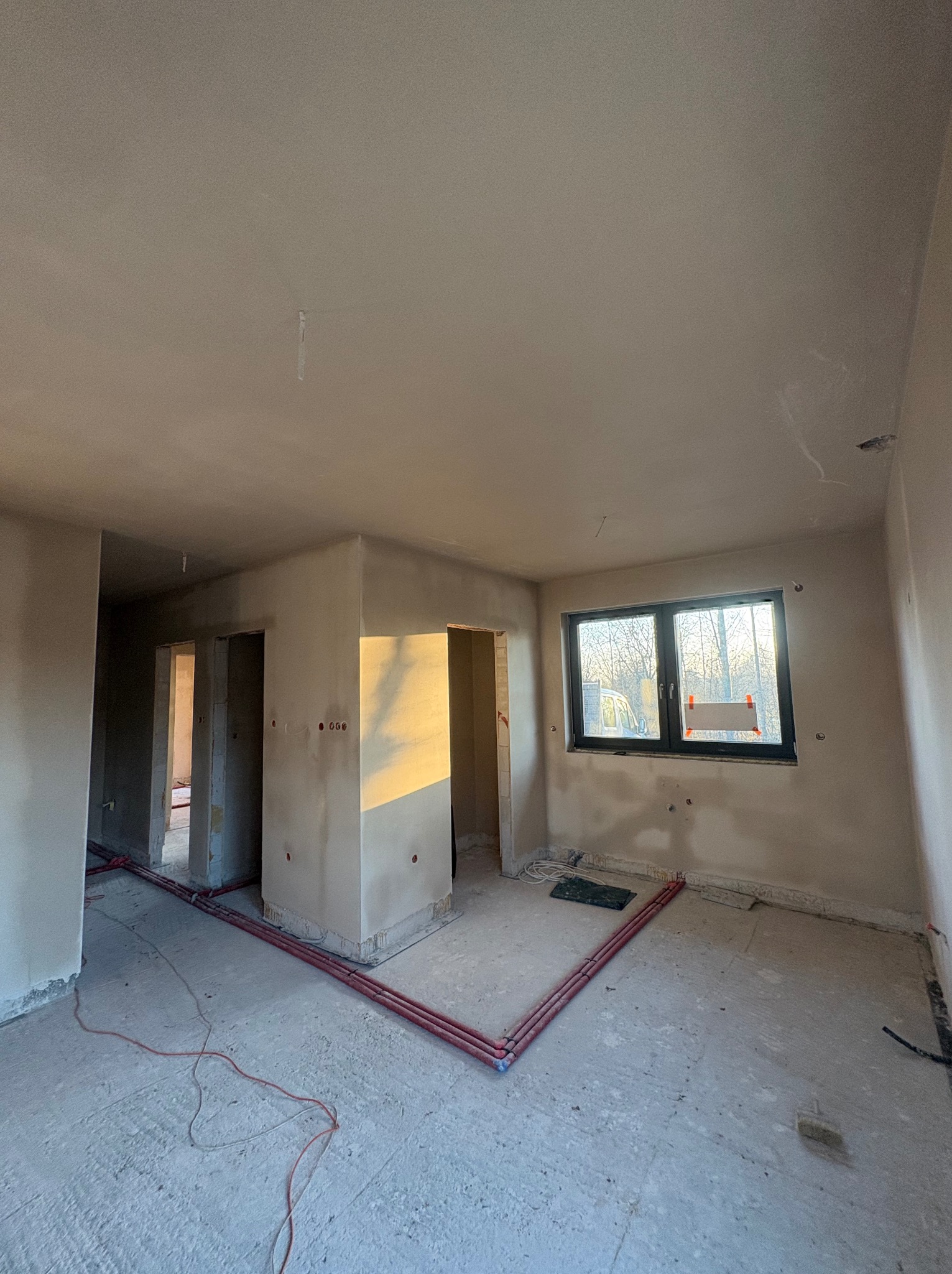 Wnętrze w trakcie remontu: ściany po gładziowaniu, widoczne instalacje podłogowe i elektryczne, okno z widokiem na drzewa. Promienie słoneczne oświetlają fragment ściany.