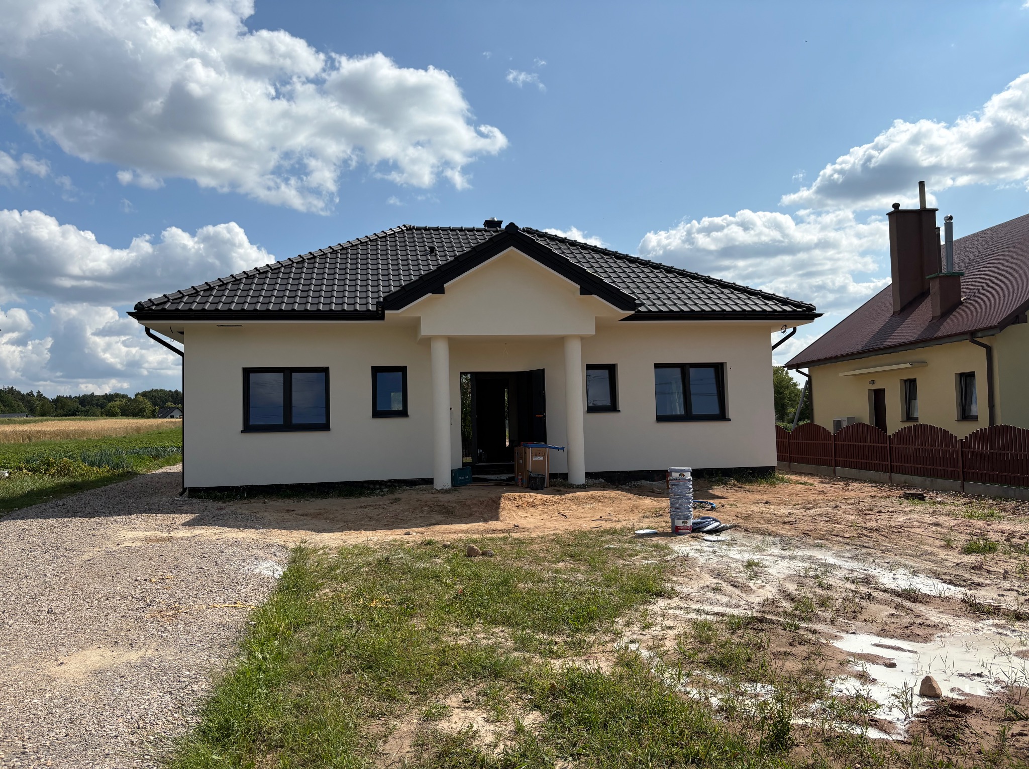 Nowoczesny, parterowy dom z białymi ścianami i ciemną dachówką, otoczony ziemią i trawą. Widoczne niebo z chmurami i fragment sąsiedniego budynku.