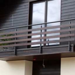 SYNCTERRA SPÓŁKA Z OGRANICZONĄ ODPOWIEDZIALNOŚCIĄ - Nowoczesny balkon z drewnianą balustradą i metalowymi elementami, dopasowany do ciemnej elewacji domu. Widok z dołu, podkreślający solidną konstrukcję.