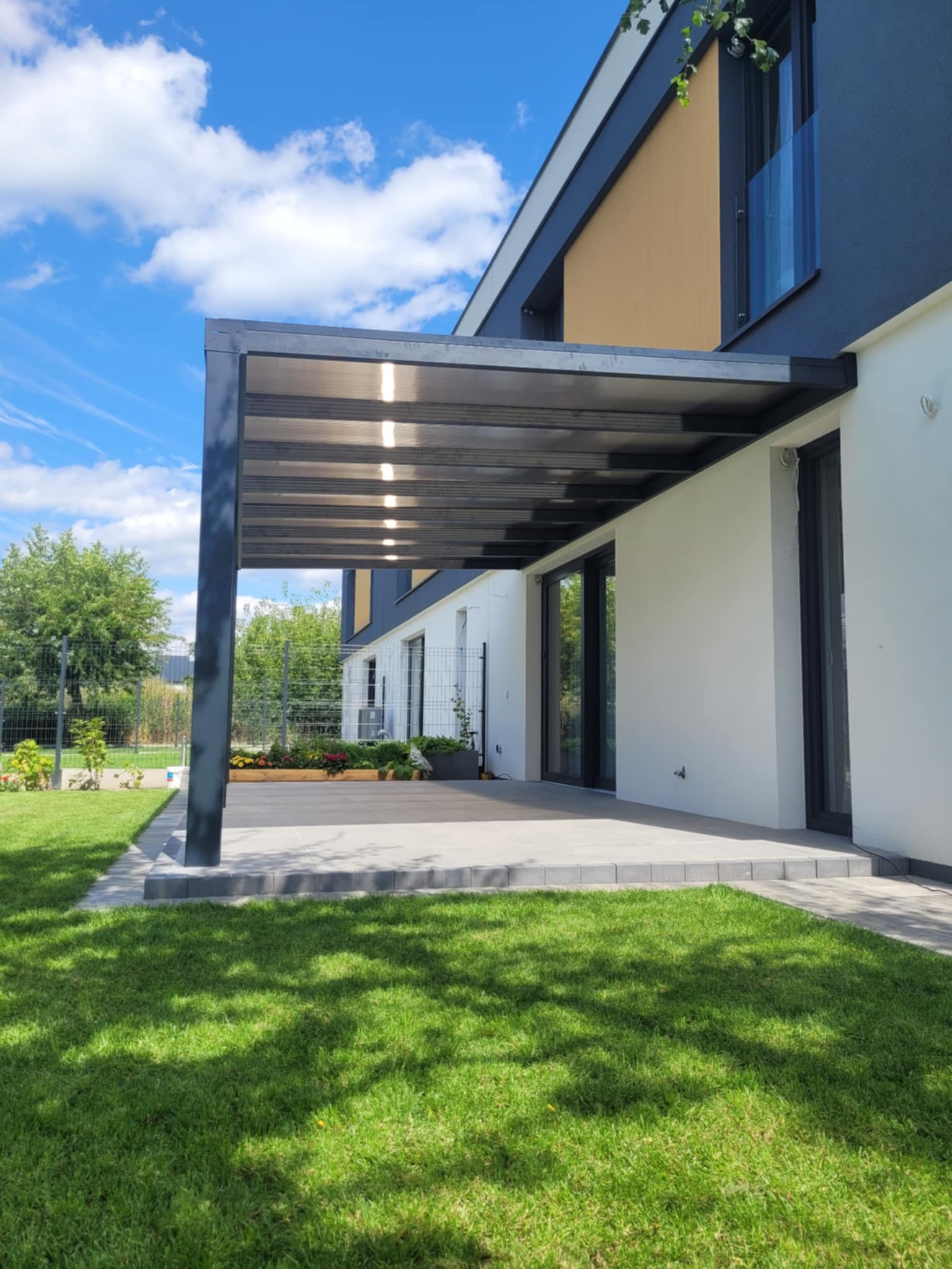 Nowoczesna pergola z ciemną konstrukcją i przezroczystym zadaszeniem, przylegająca do białego budynku z dużymi oknami, otoczona zielonym trawnikiem i ogrodzeniem.