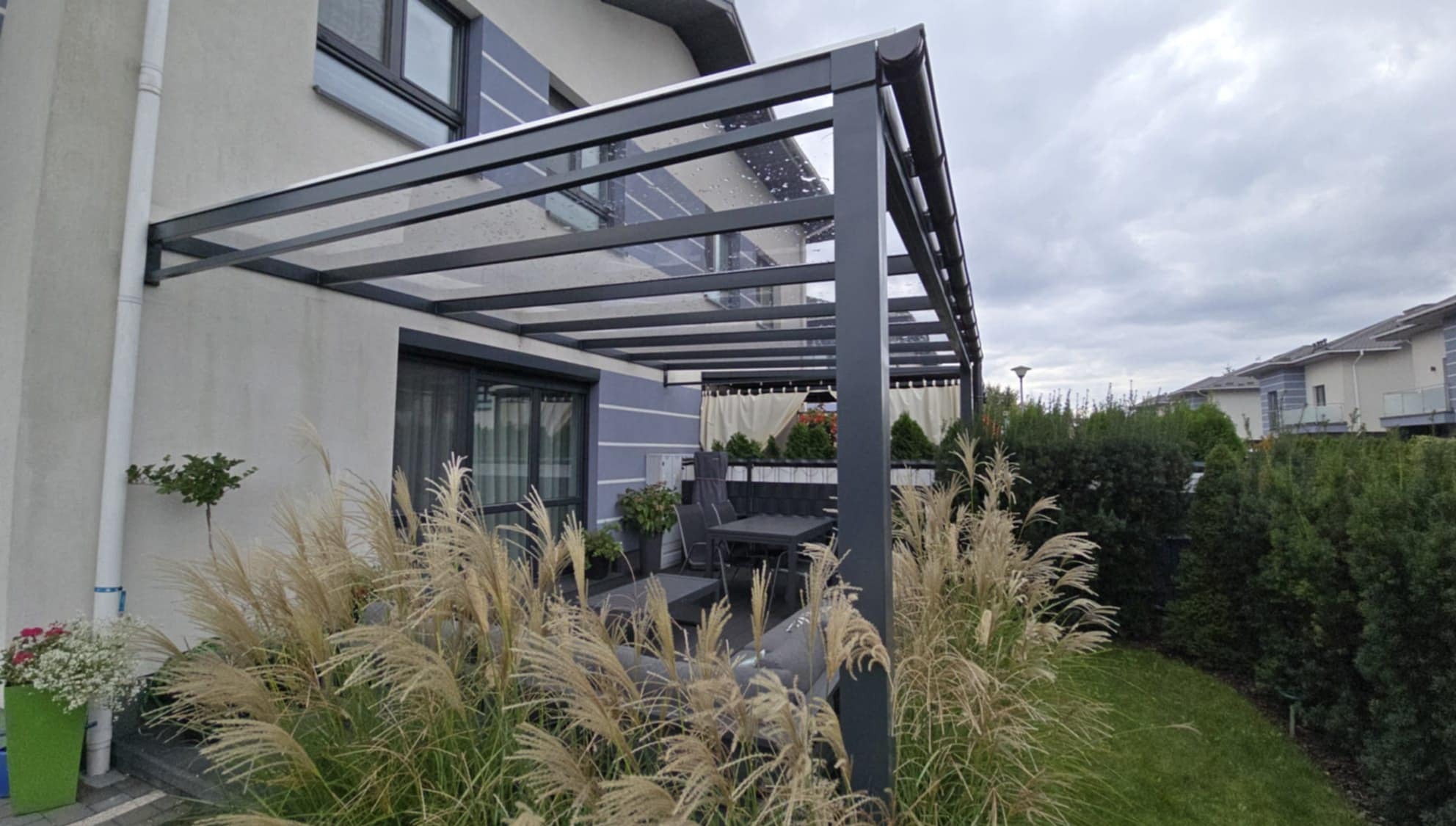 Nowoczesna pergola metalowa z przezroczystym zadaszeniem, osłaniająca taras z meblami ogrodowymi, otoczona ozdobnymi trawami i zielenią. Widok na dom w tle.