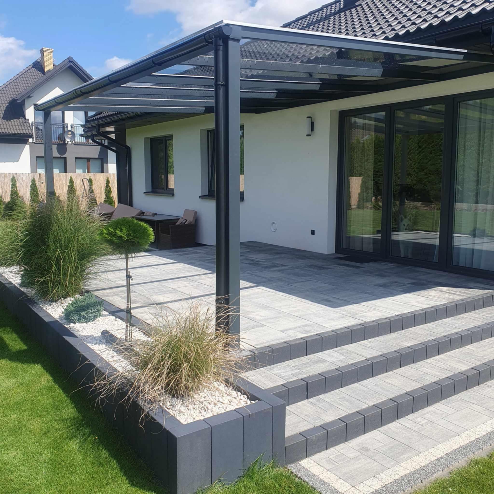 Nowoczesna pergola z metalową konstrukcją i szklanym dachem, osłaniająca taras z kostki brukowej. Widoczne elementy architektury ogrodowej i domu.