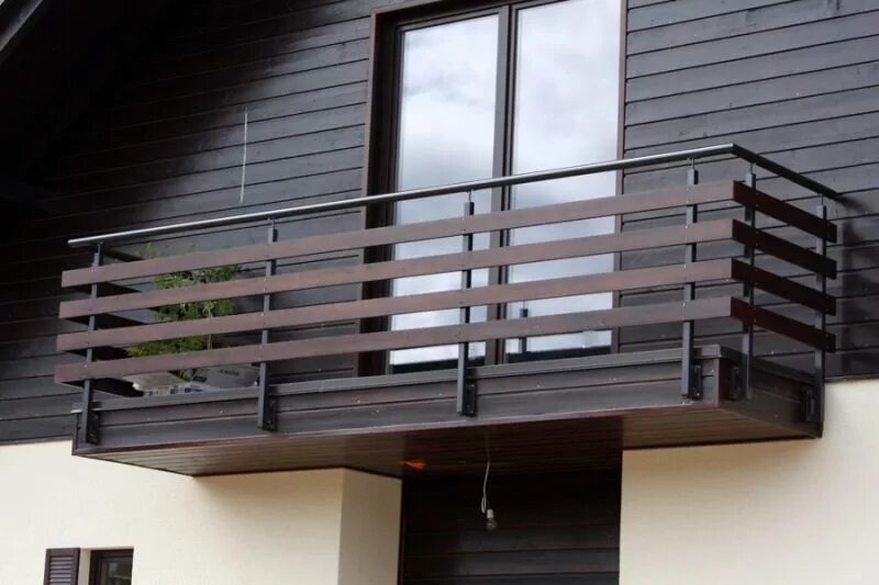 Nowoczesny balkon z drewnianą balustradą i metalowymi elementami, dopasowany do ciemnej elewacji domu. Widok z dołu, podkreślający solidną konstrukcję.