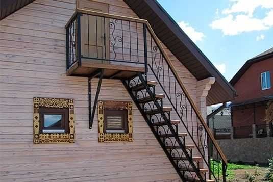Metalowe schody zewnętrzne z ozdobnymi balustradami prowadzące na balkon w drewnianym domu, Warszawa. Konstrukcja wsparta na metalowych wspornikach.