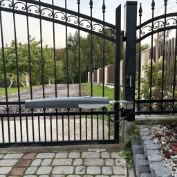 AK ENERGY Arkadiusz Krzywda - Czarna, metalowa brama z zamontowaną automatyką, widoczna z bliska na tle zieleni ogrodu i kostki brukowej. Napęd bramy w kolorze szarym.