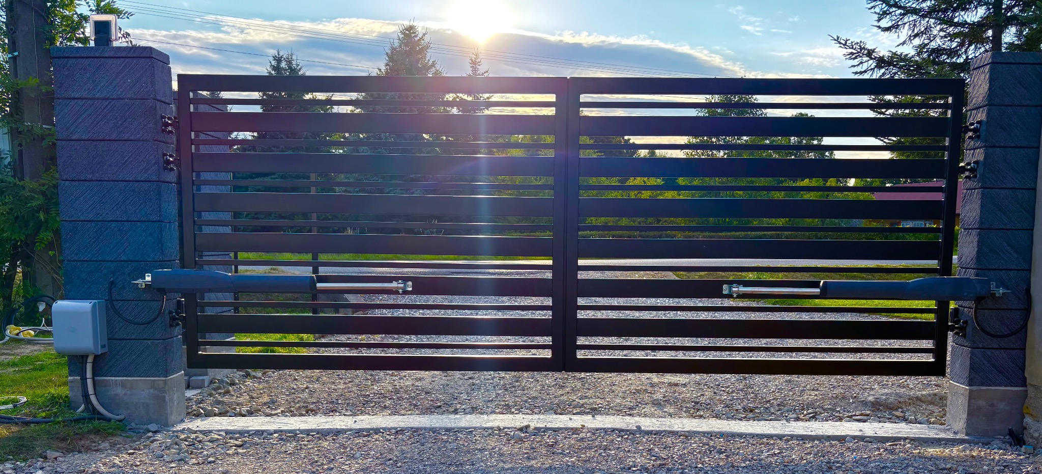Nowoczesna, pozioma brama wjazdowa z automatyką, zamontowana na kamiennych słupkach. Widoczne siłowniki i skrzynka sterownicza. Tło: zieleń drzew i błękitne niebo.