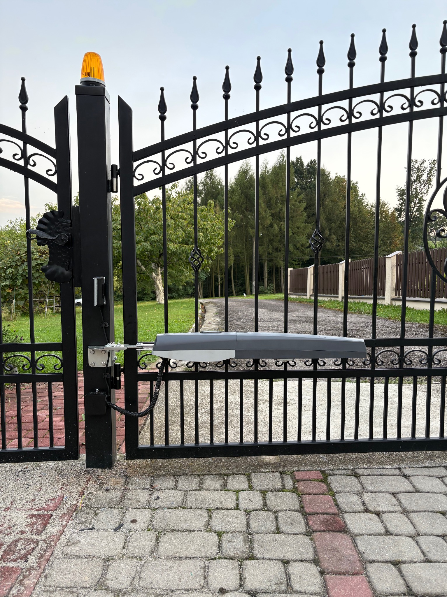 Czarna, ozdobna brama wjazdowa z zamontowaną automatyką i lampą ostrzegawczą. Widok na posesję z brukowanym podjazdem i zielenią w tle.