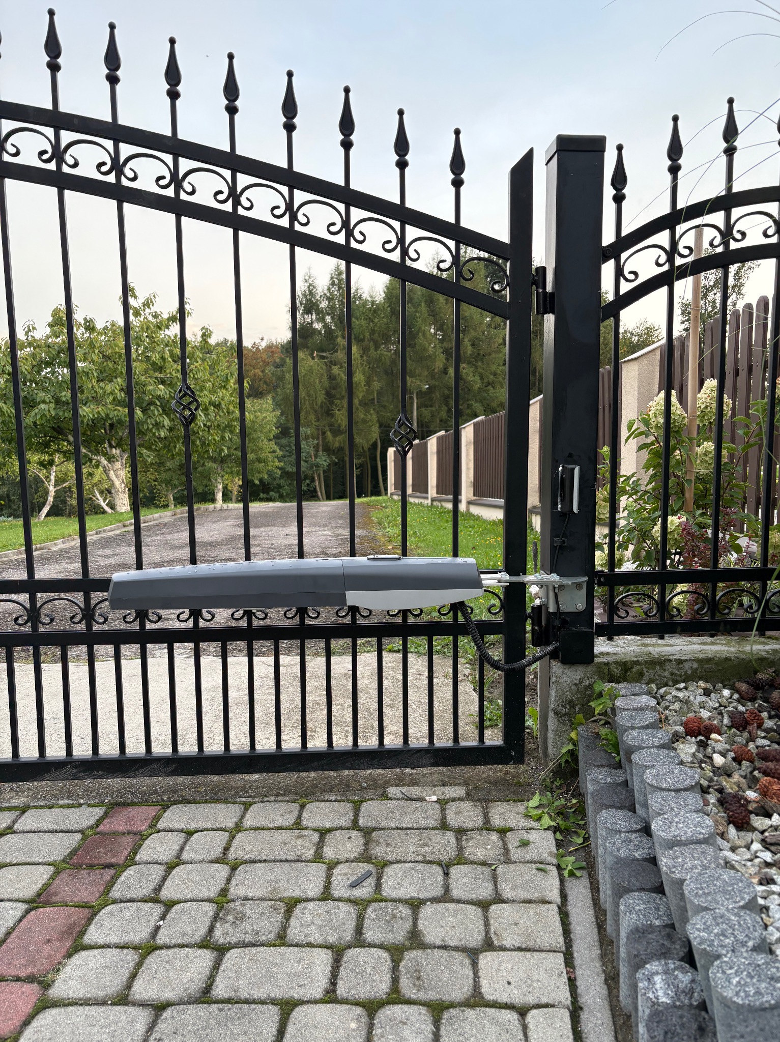 Czarna, metalowa brama z zamontowaną automatyką, widoczna z bliska na tle zieleni ogrodu i kostki brukowej. Napęd bramy w kolorze szarym.