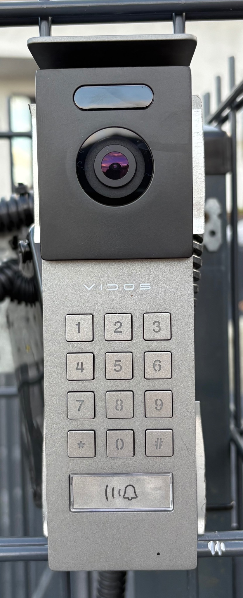 Szary panel wideodomofonu Vidos z klawiaturą numeryczną i kamerą, zamontowany na metalowym ogrodzeniu. Widoczne odbicie w obiektywie kamery.