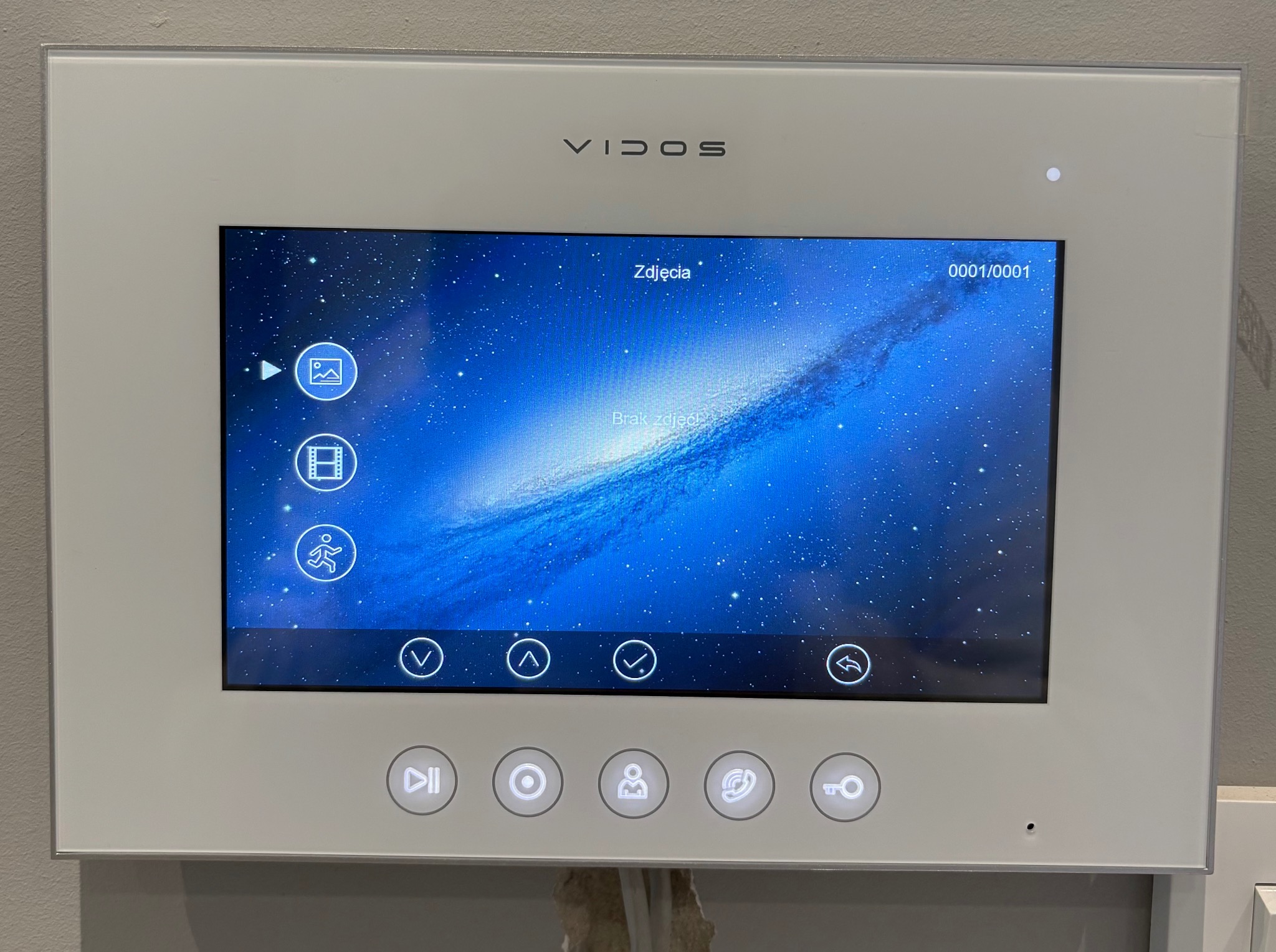 Biały panel dotykowy wideodomofonu Vidos z interfejsem użytkownika wyświetlającym galaktykę. Widoczne ikony i przyciski funkcyjne.