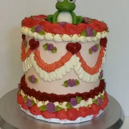 Tort w stylu retro z r&oacute;żową polewą, dekoracjami z kremu i figurką żaby w koronie na wierzchu, umieszczony na srebrnej paterze.