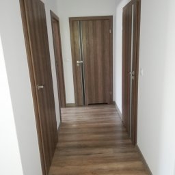 USŁUGI BUDOWLANE ADRIAN KONTOWSKI - Korytarz z trzema parami drewnianych drzwi i panelami podłogowymi w odcieniach brązu. Widok w perspektywie, ściany białe, minimalistyczny styl.