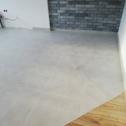 USŁUGI BUDOWLANE ADRIAN KONTOWSKI - Jasnoszare płytki podłogowe w nowym wnętrzu, z fragmentem drewnianej podłogi i dekoracyjną ścianą z szarej cegły. Widoczne gniazdka elektryczne.
