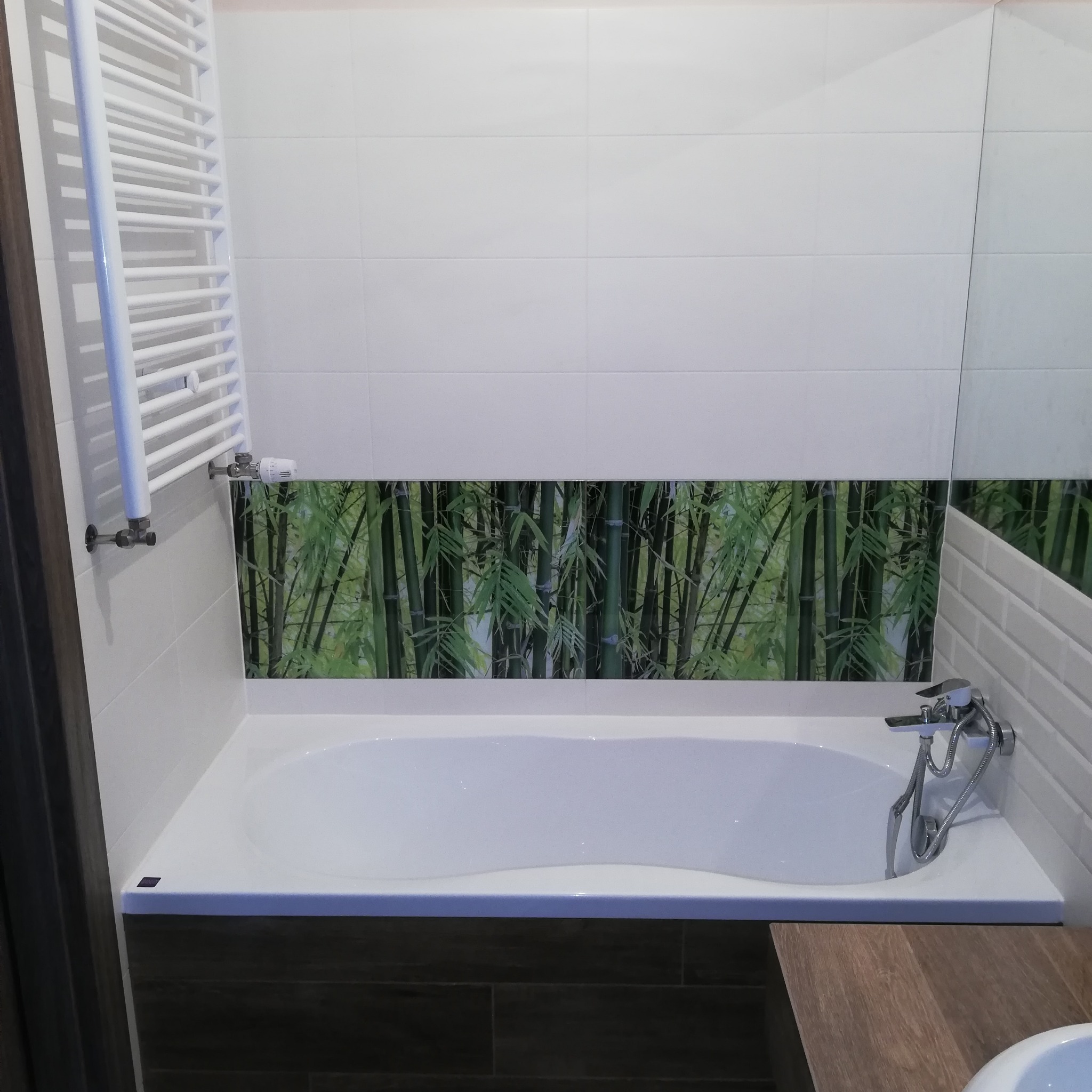 Nowoczesna łazienka z białą wanną, grzejnikiem drabinkowym i płytkami z motywem bambusa. Ujęcie na fragment łazienki z bliska, widoczne detale wykończenia.