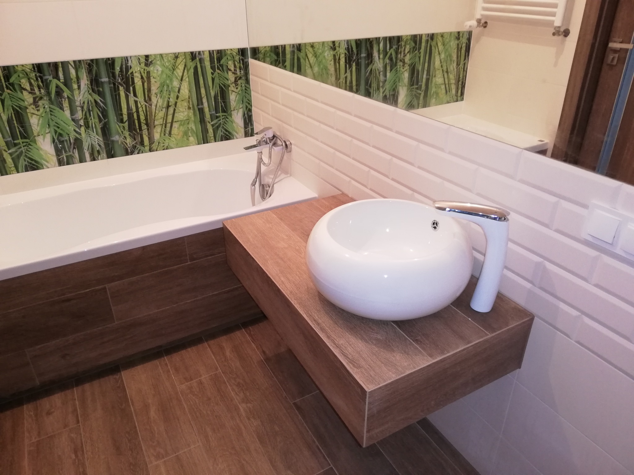 Nowoczesna łazienka z białą umywalką na drewnianym blacie, białe płytki imitujące cegłę i dekoracyjny panel z motywem bambusa nad wanną.