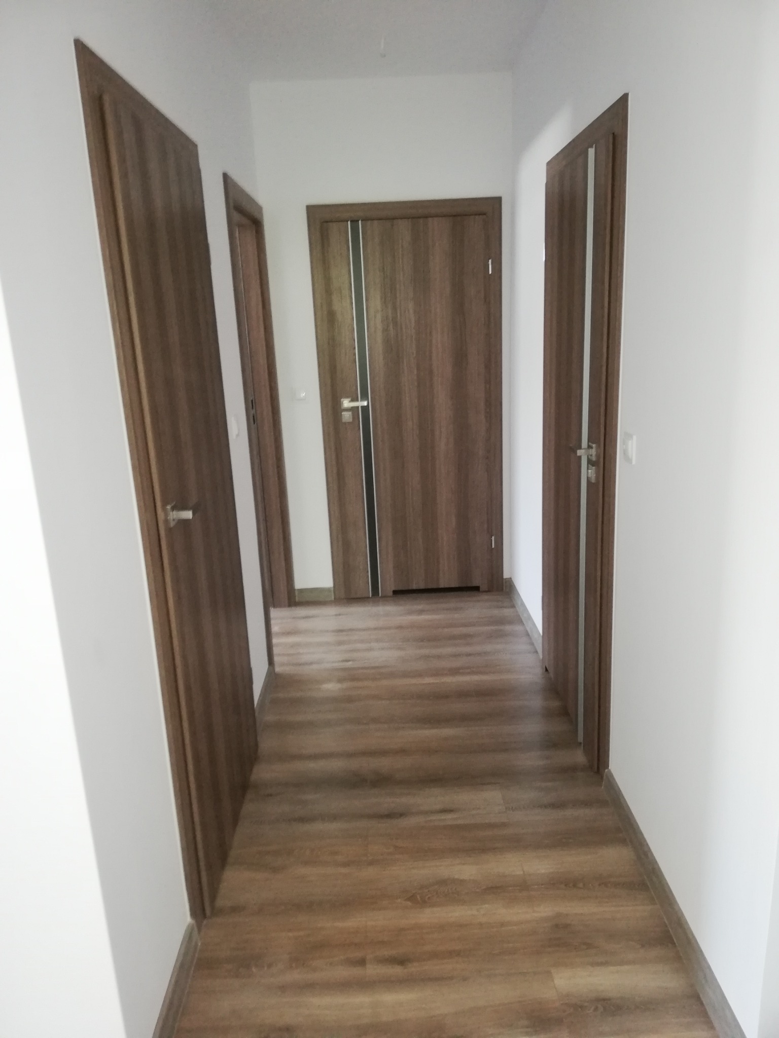 Korytarz z trzema parami drewnianych drzwi i panelami podłogowymi w odcieniach brązu. Widok w perspektywie, ściany białe, minimalistyczny styl.