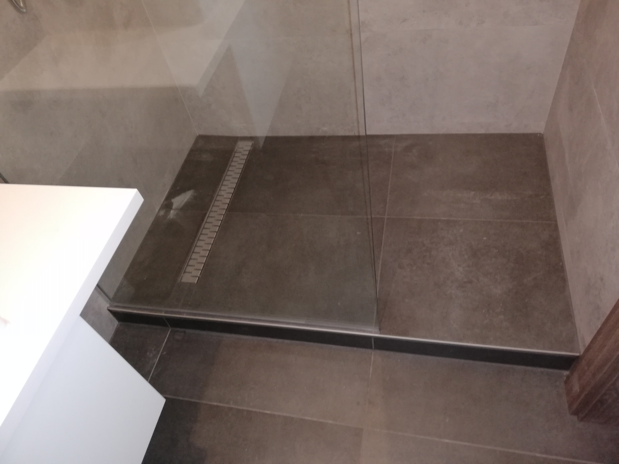 Nowoczesna kabina prysznicowa z odpływem liniowym i szarymi płytkami. Minimalistyczny design, widoczny fragment umywalki. Wykończenie łazienki w stonowanych barwach.