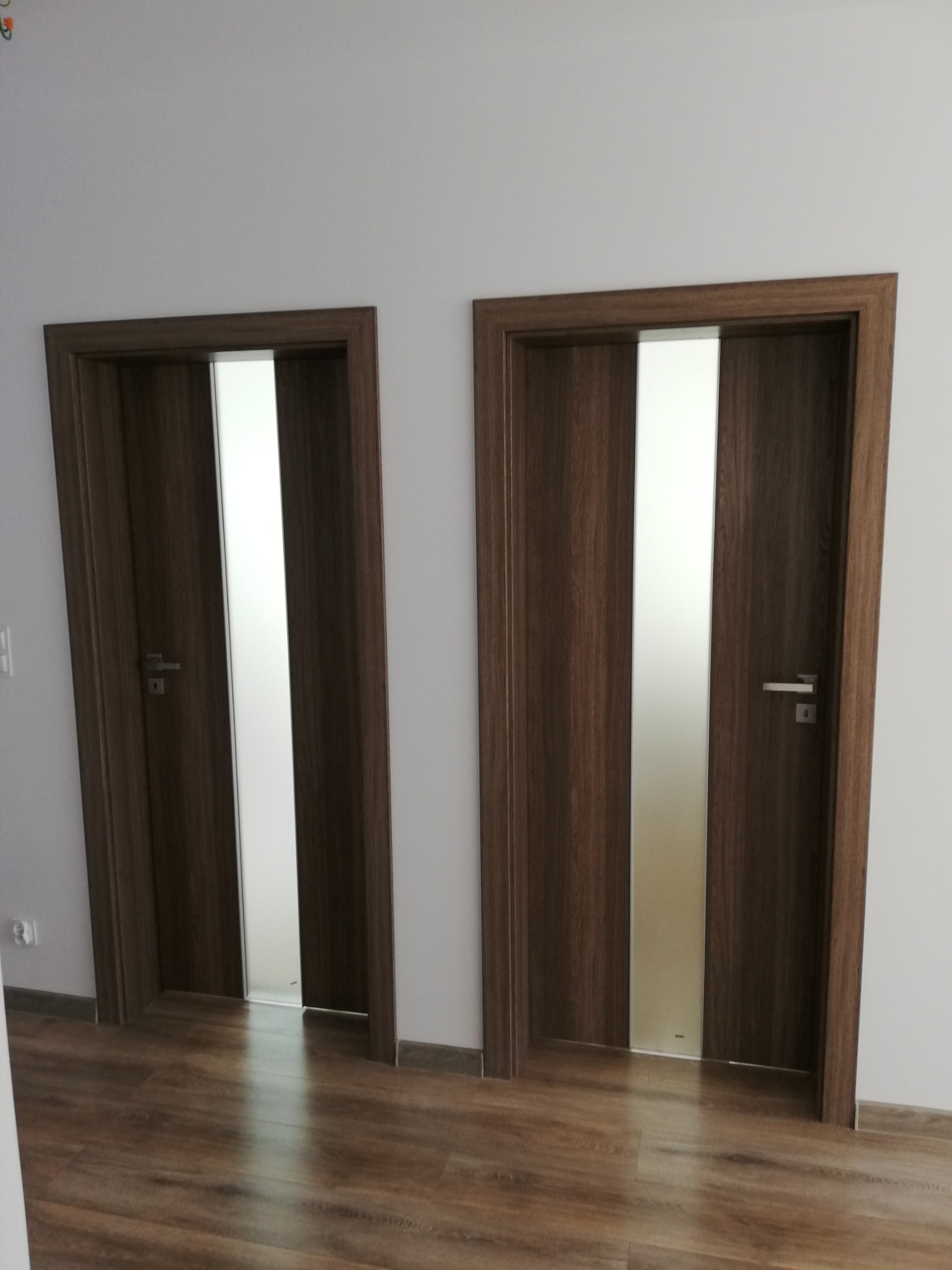 Dwa identyczne, brązowe drzwi wewnętrzne z matowym, pionowym przeszkleniem, zamontowane w jasnym pomieszczeniu z panelami podłogowymi imitującymi drewno.