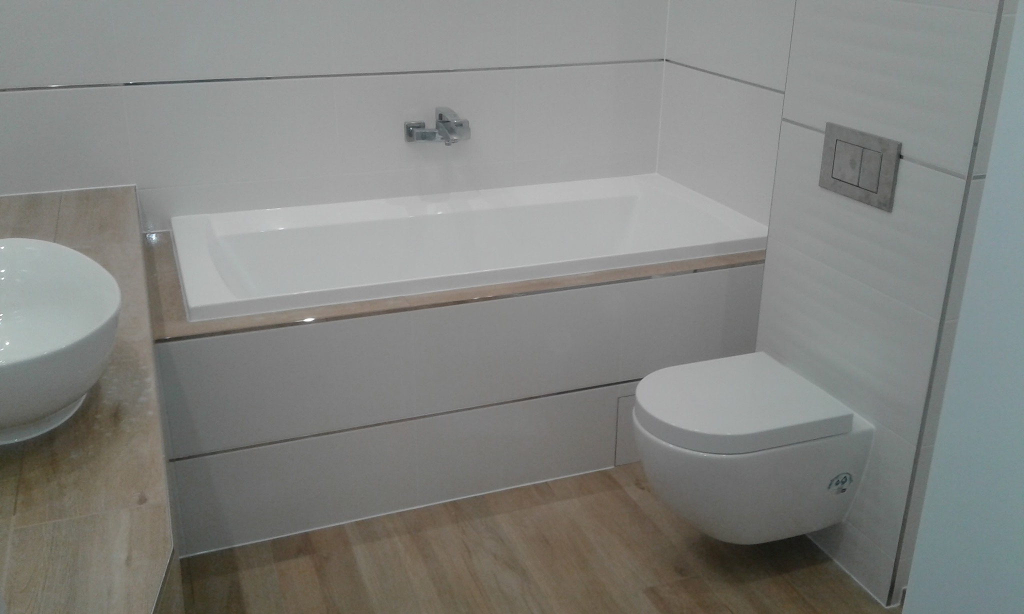 Nowoczesna łazienka z białą wanną, podwieszaną toaletą i umywalką nablatową. Jasne płytki i drewnopodobna podłoga tworzą minimalistyczny, elegancki wystrój.