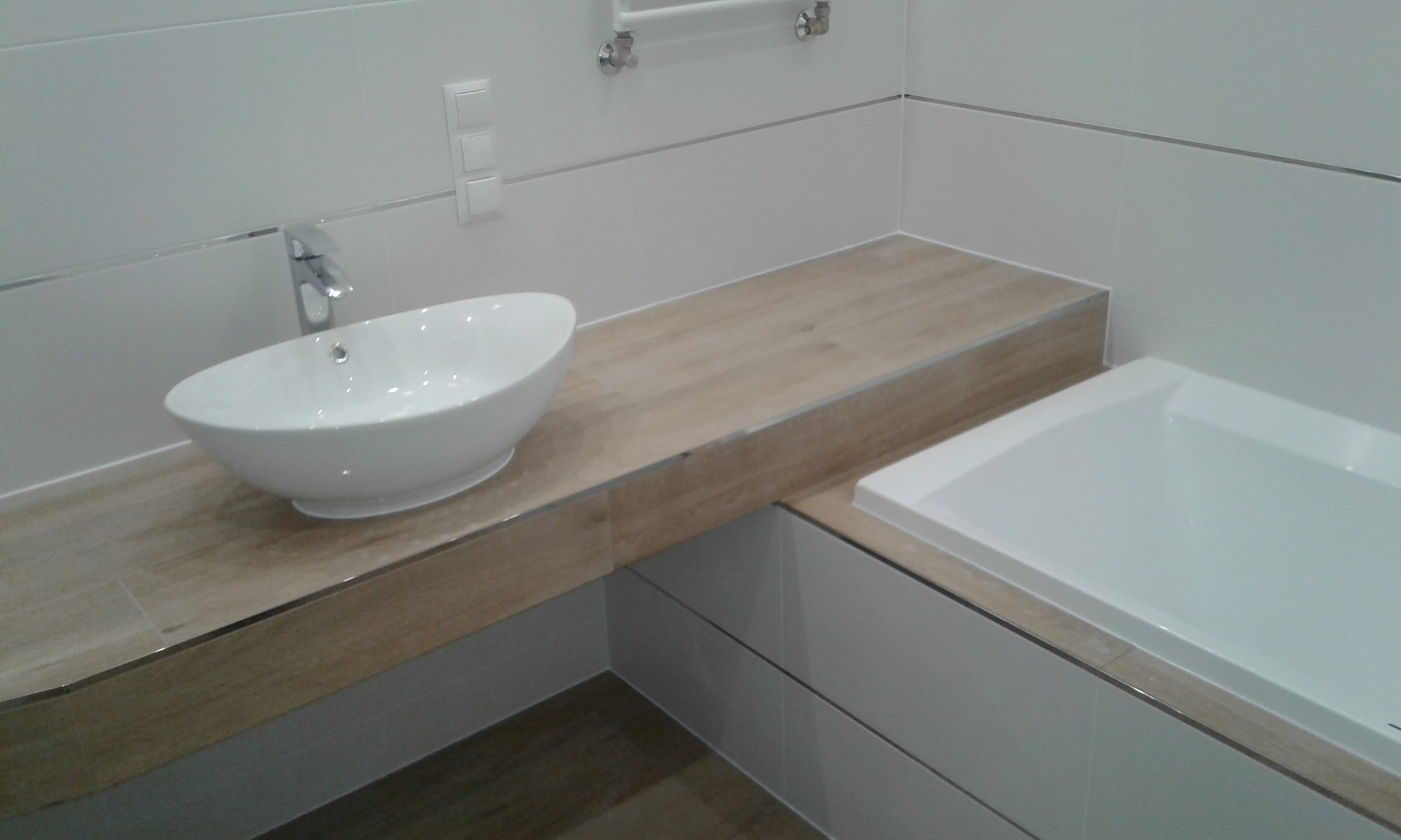 Nowoczesna łazienka z owalną umywalką nablatową, drewnopodobnym blatem i białą wanną. Minimalistyczne płytki i chromowane detale.