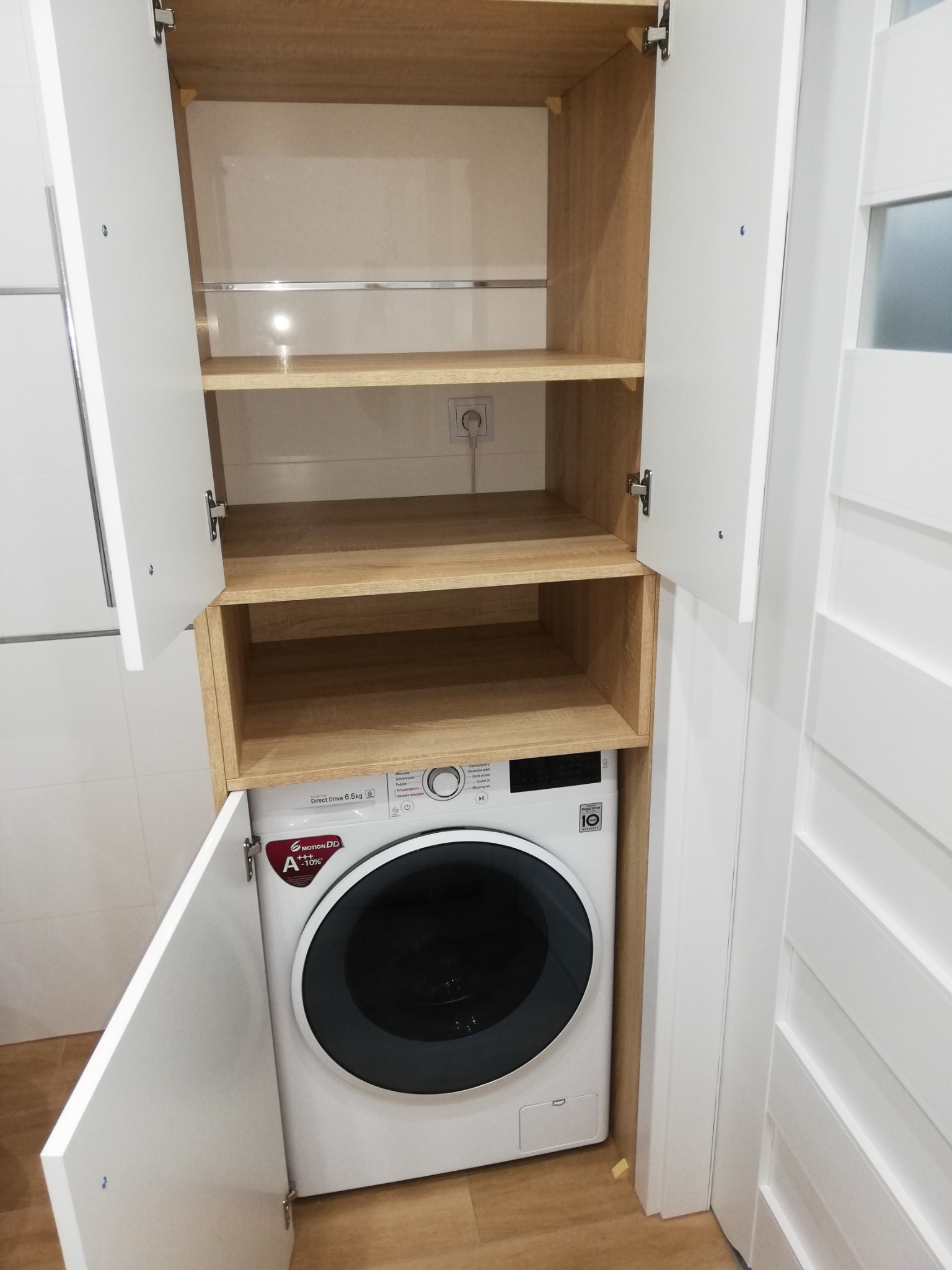 Funkcjonalna zabudowa pralki z półkami w łazience w Goleniowie. Białe fronty szafek i drewnopodobne półki tworzą spójną całość. Widoczna pralka LG.
